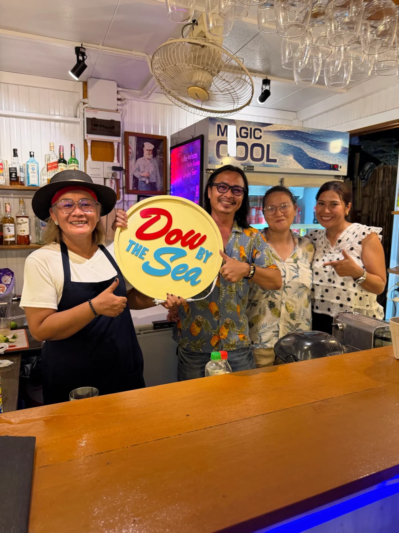 จากซ้ายไปขวา: Dow, Jek, Faa และ Anne ยืนด้วยกันหลังบาร์พร้อมป้าย Dow by the sea